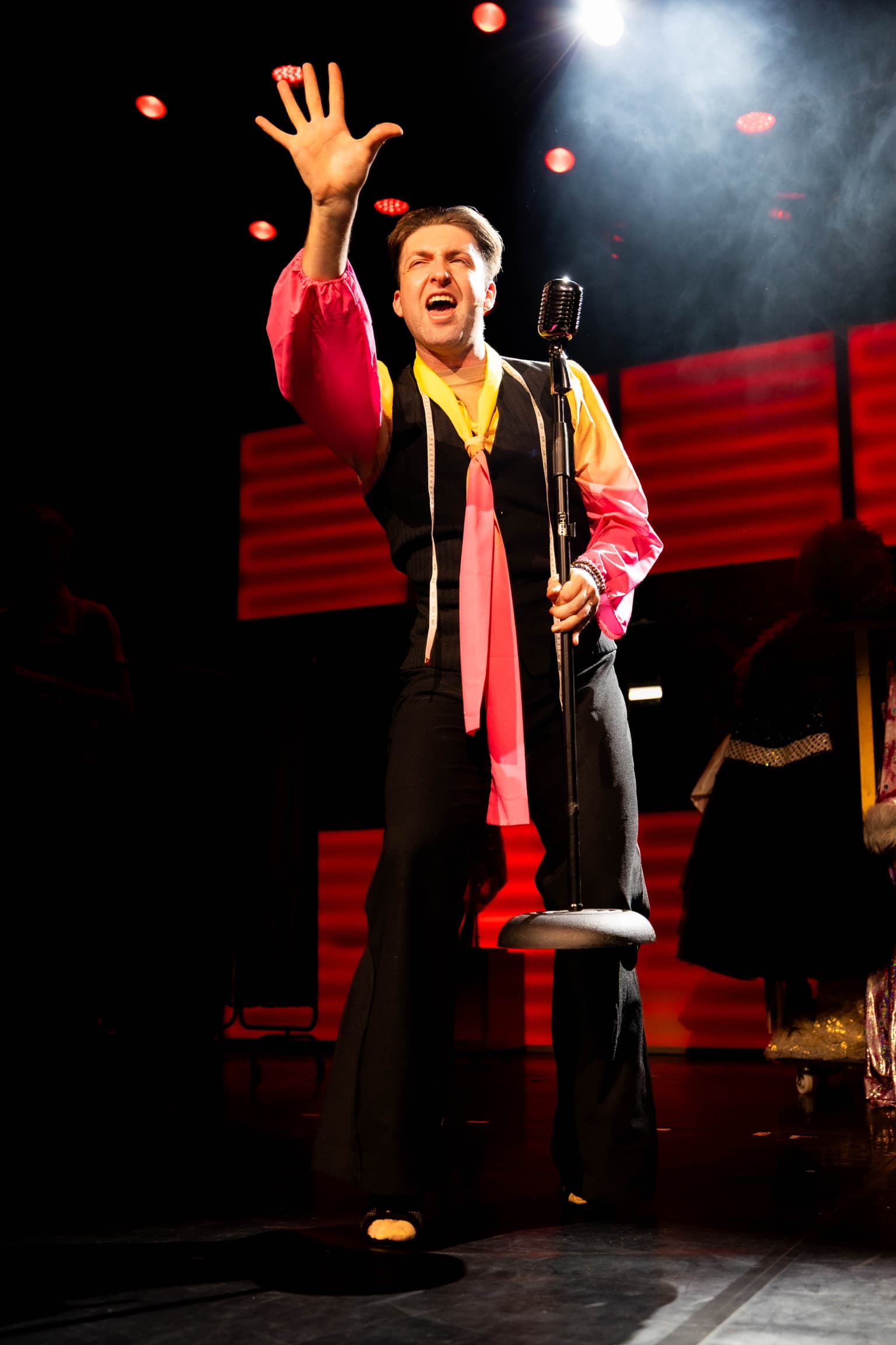 Hugo poses towards the audience while singing. He holds an old-style microphone on a stand. The LED wall is lit up red.