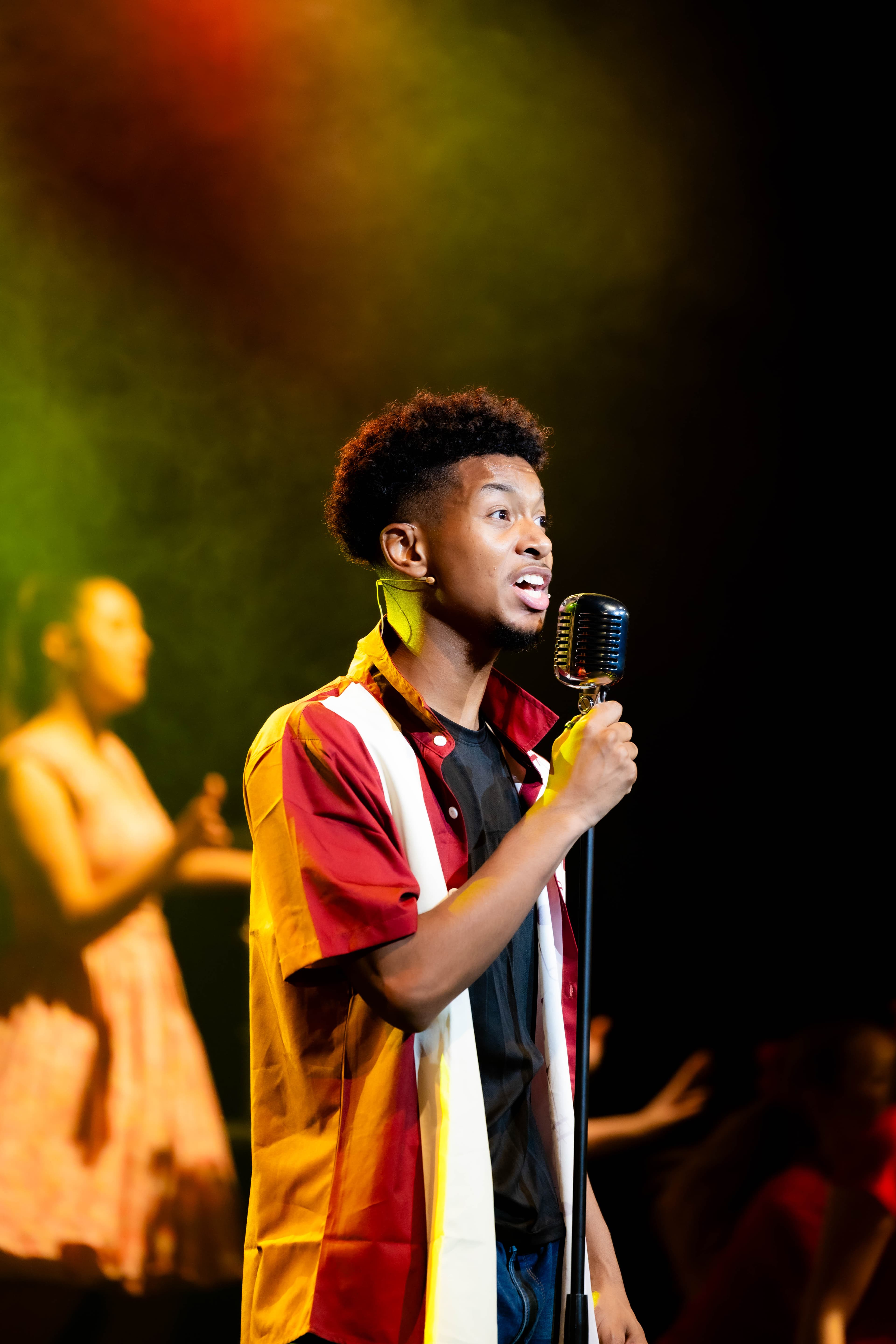 Doody stands and sings a solo song into an old-style microphone on a stand. He is gently lit from behind in a yellow and red spotlight.