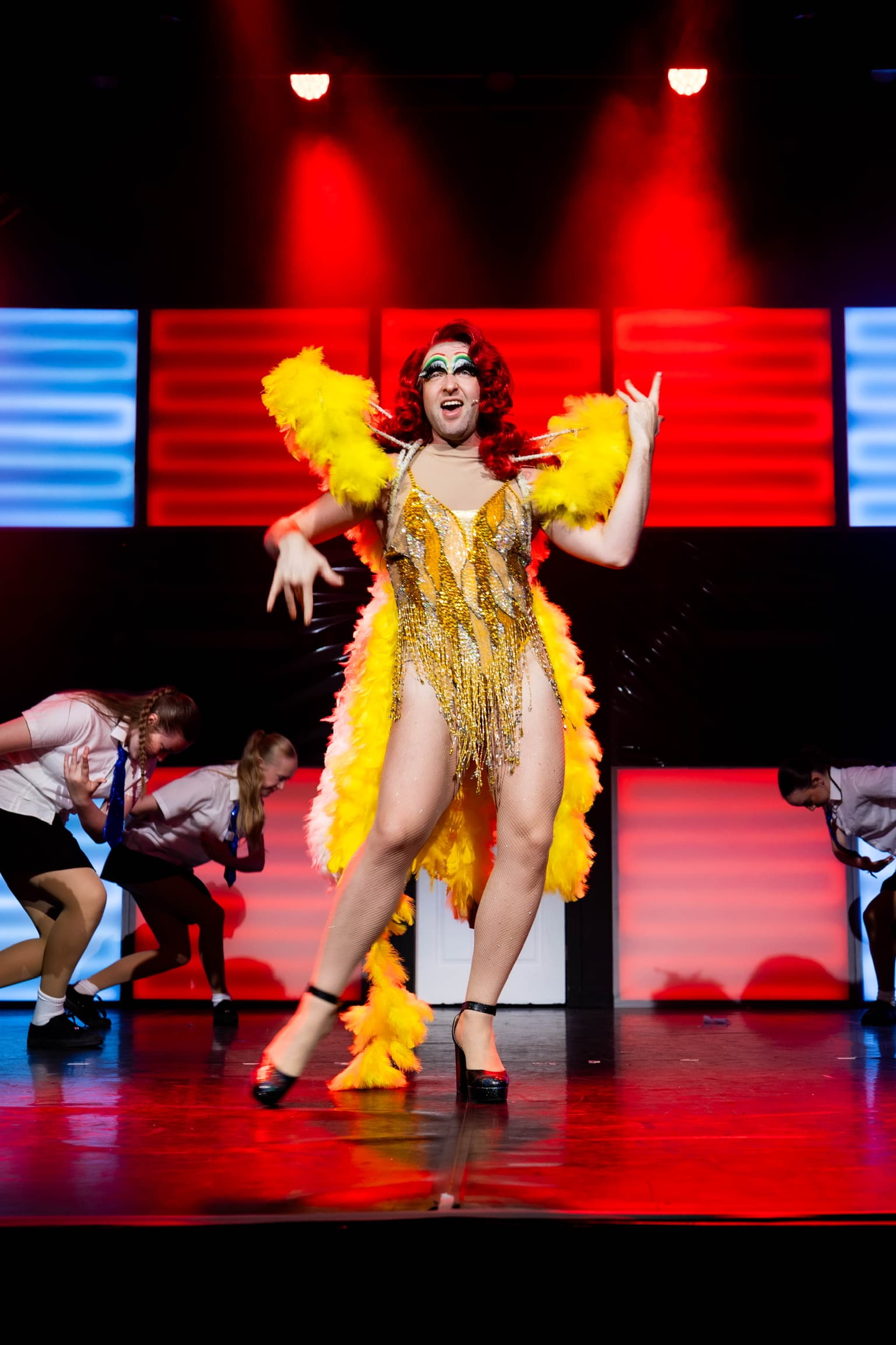 Loco Chanelle poses on stage in an extravagant yellow drag dress, with bright red hair. She is spotlight by red lights. The LED wall is lit up Red and White.