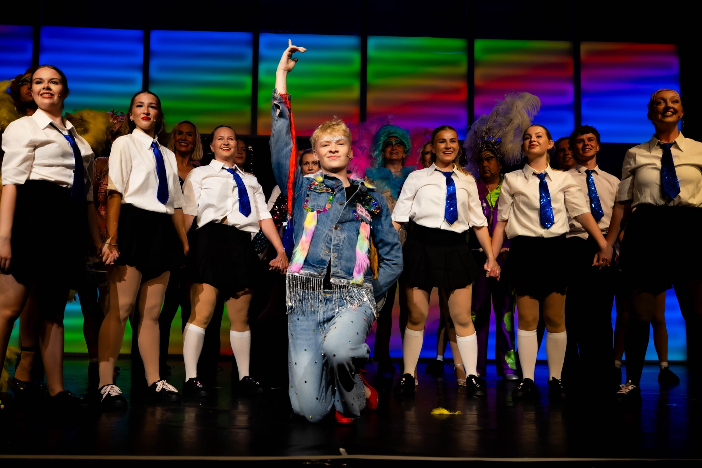 Jamie poses on one knee with one arm in the air at the front of the stage. He is surrounded by students in shirts and sparkly blue ties. The LED wall is lit up rainbow.