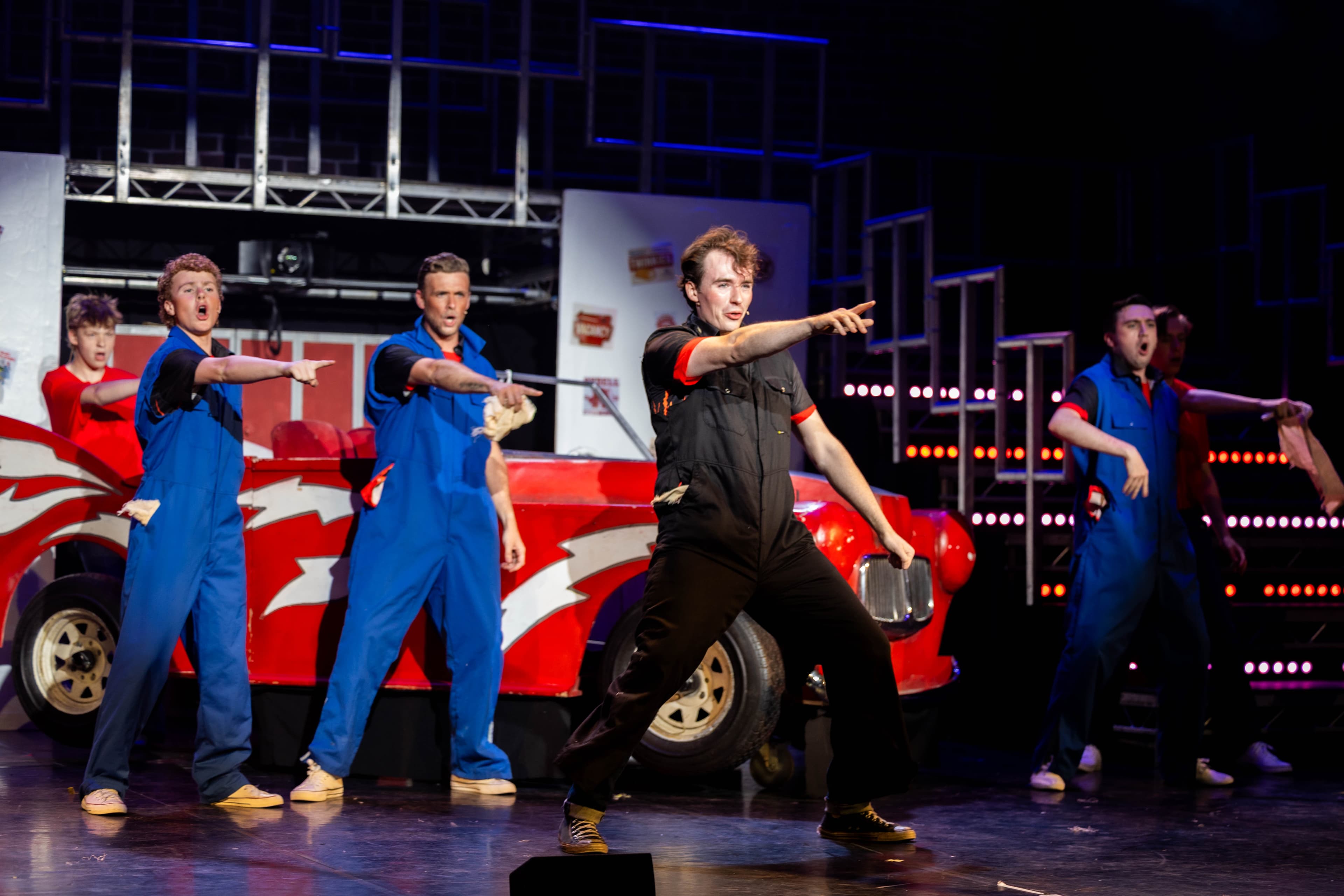 The T-Birds dance in front of the Greased Lightning car in mechanics boiler suits. They stand with their arms pointing out to the audience.