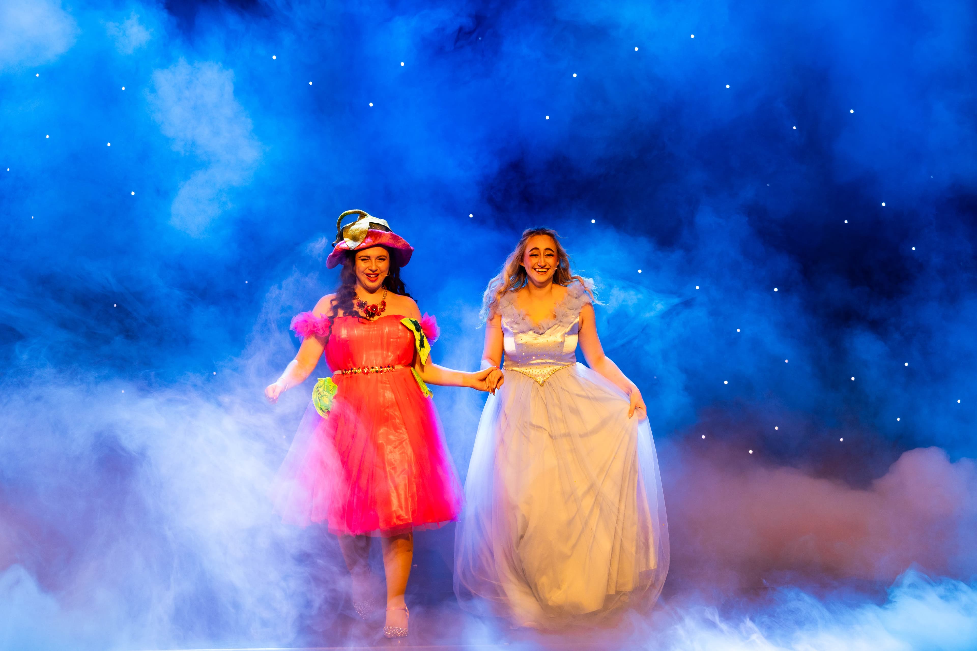 The Fairy Godmother and Cinderella emerge from a thick cloud of smoke after Cinderella transforms into her beautiful ball gown. The stage is lit blue and stars sparkle behind them.