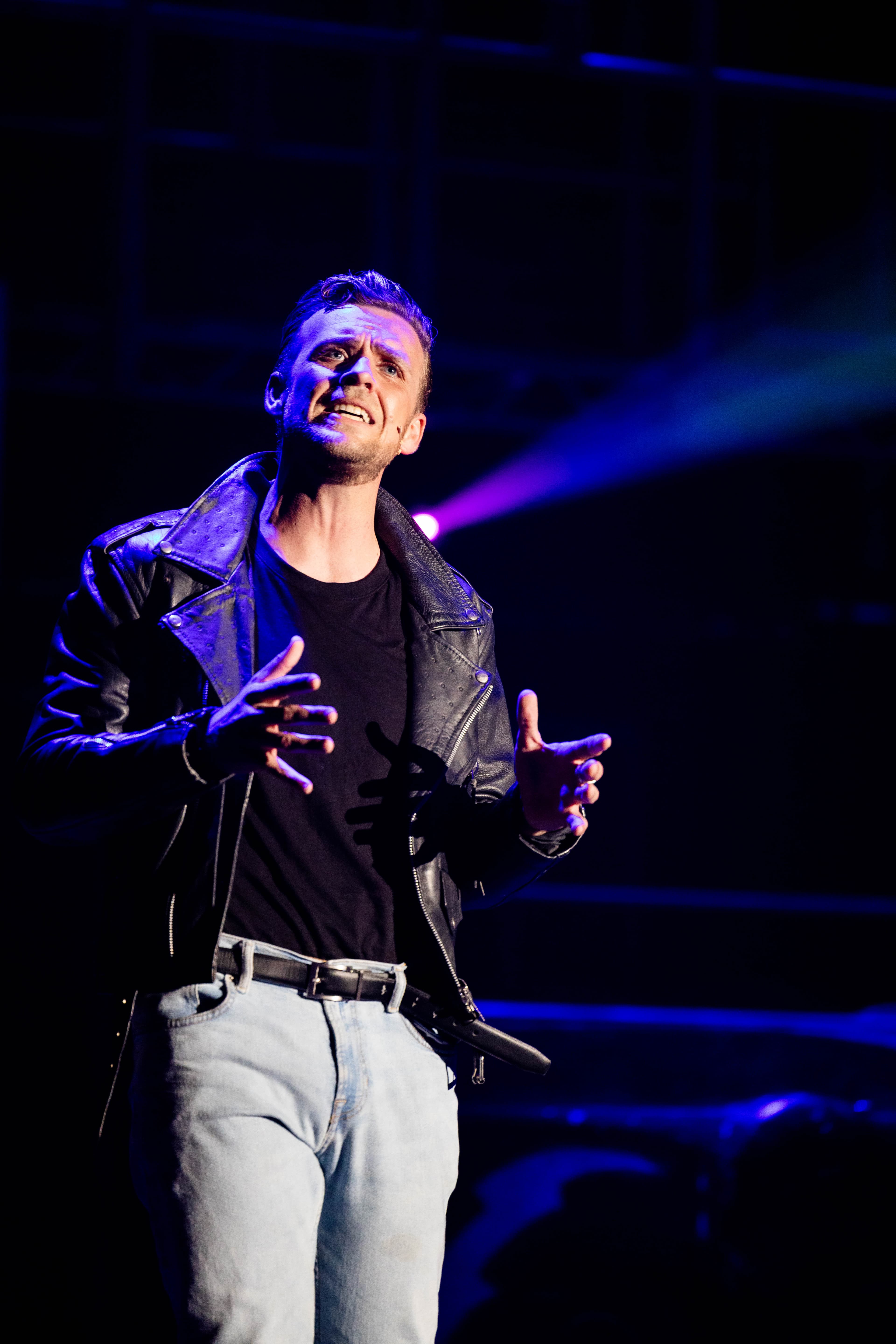 Danny Zuko poses while singing a song. Over his shoulder is the beam of a projector shining through the haze.