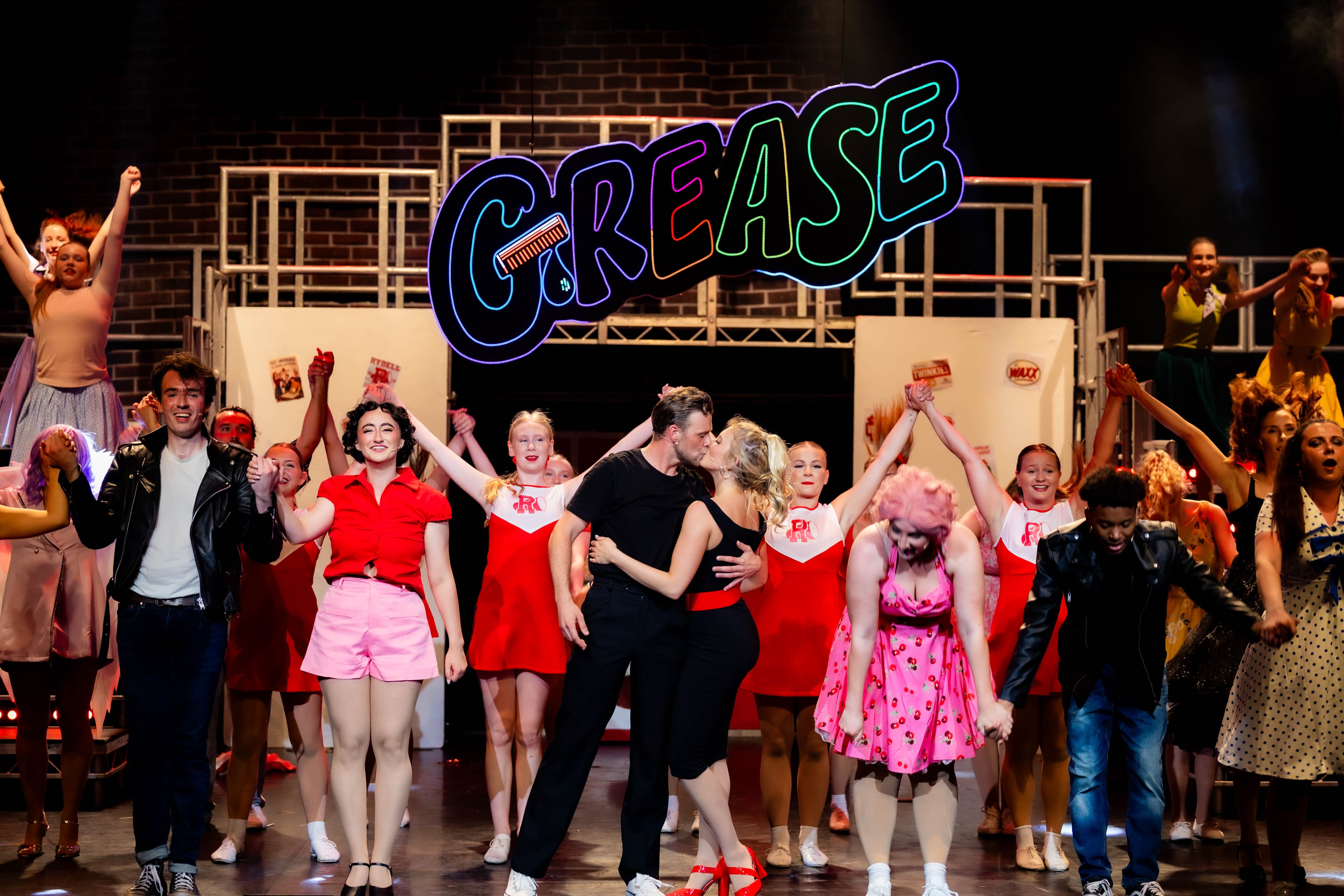 Danny and Sandy kiss under a LED Neon Grease sign, lit up in a rainbow. They are surrounded by the rest of the cast posing.
