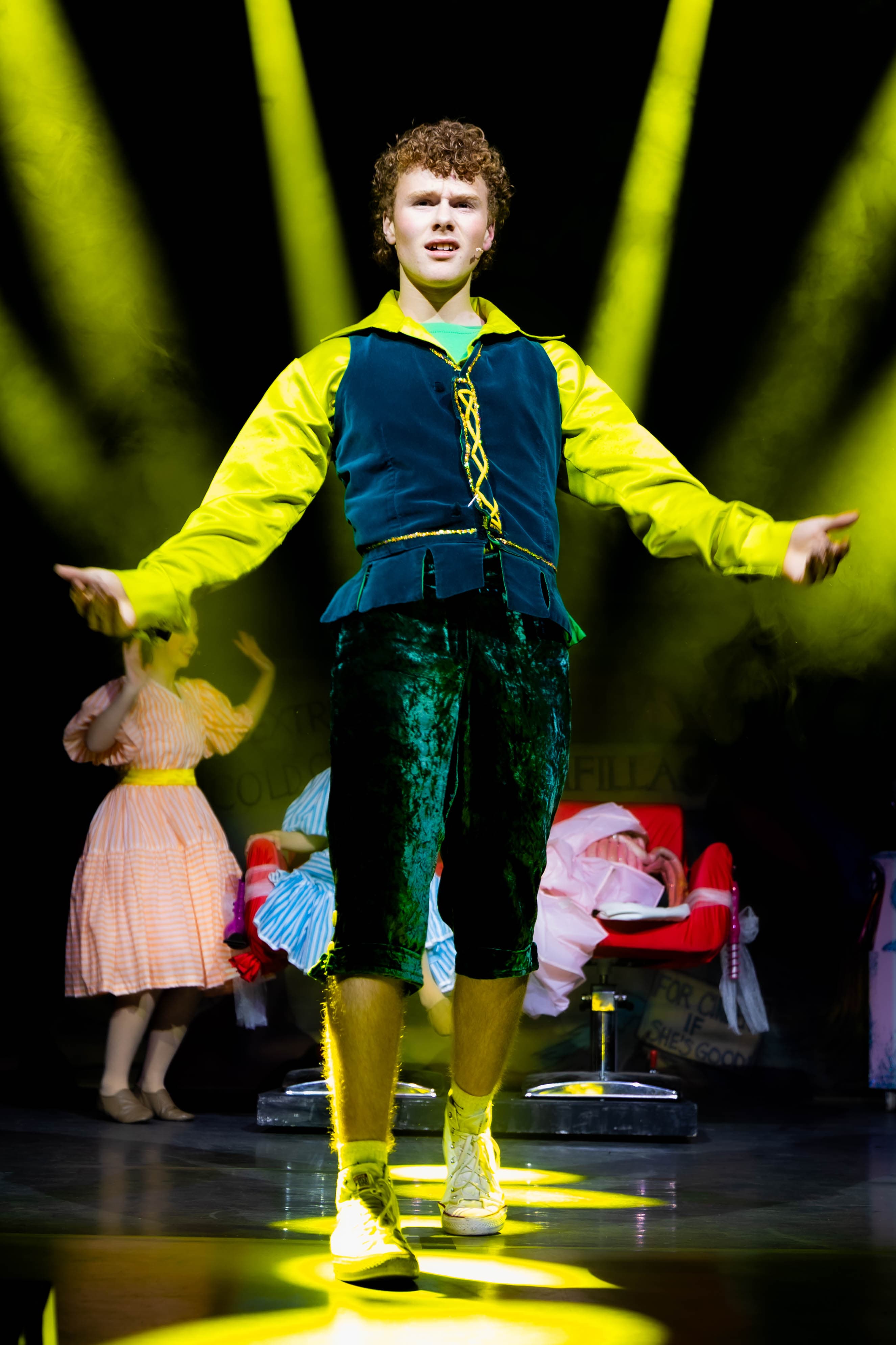 Jack poses at the front of the stage with his arms out. He is lit from above by several sharp yellow spotlights.