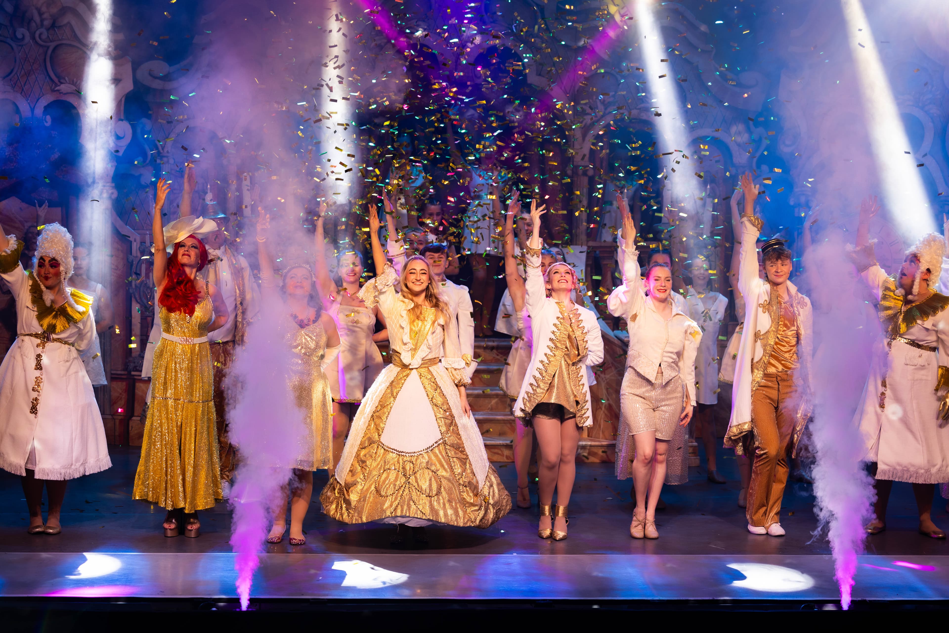 The cast of Cinderella post in ornate gold and while finale outfits. Smoke cannons fire in front of them, and gold confetti falls from above.