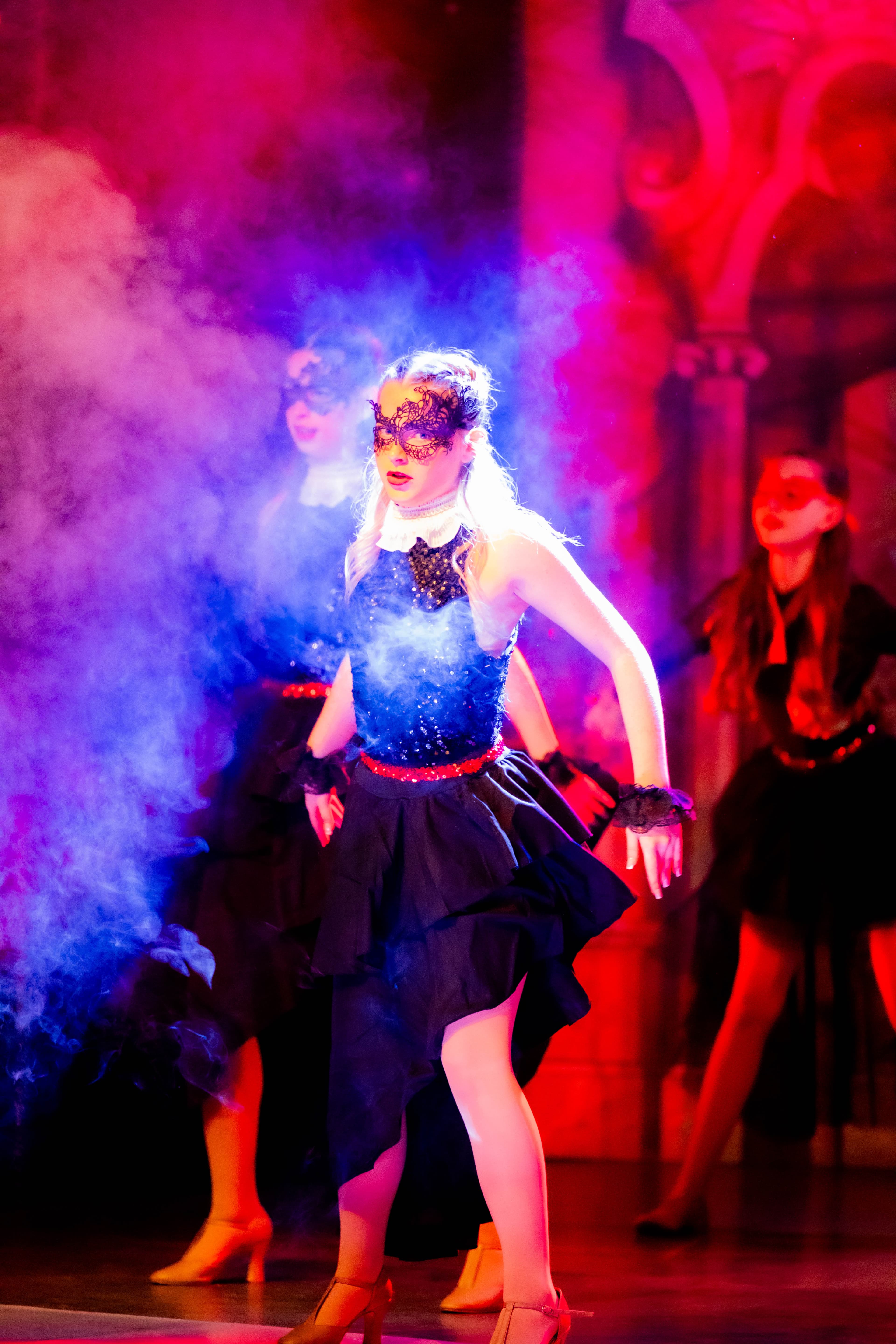 A dancer dances in a cloud of smoke. She wears a masquerade ball outfit and mask. She is lit in a white spotlight, with the stage lit red.