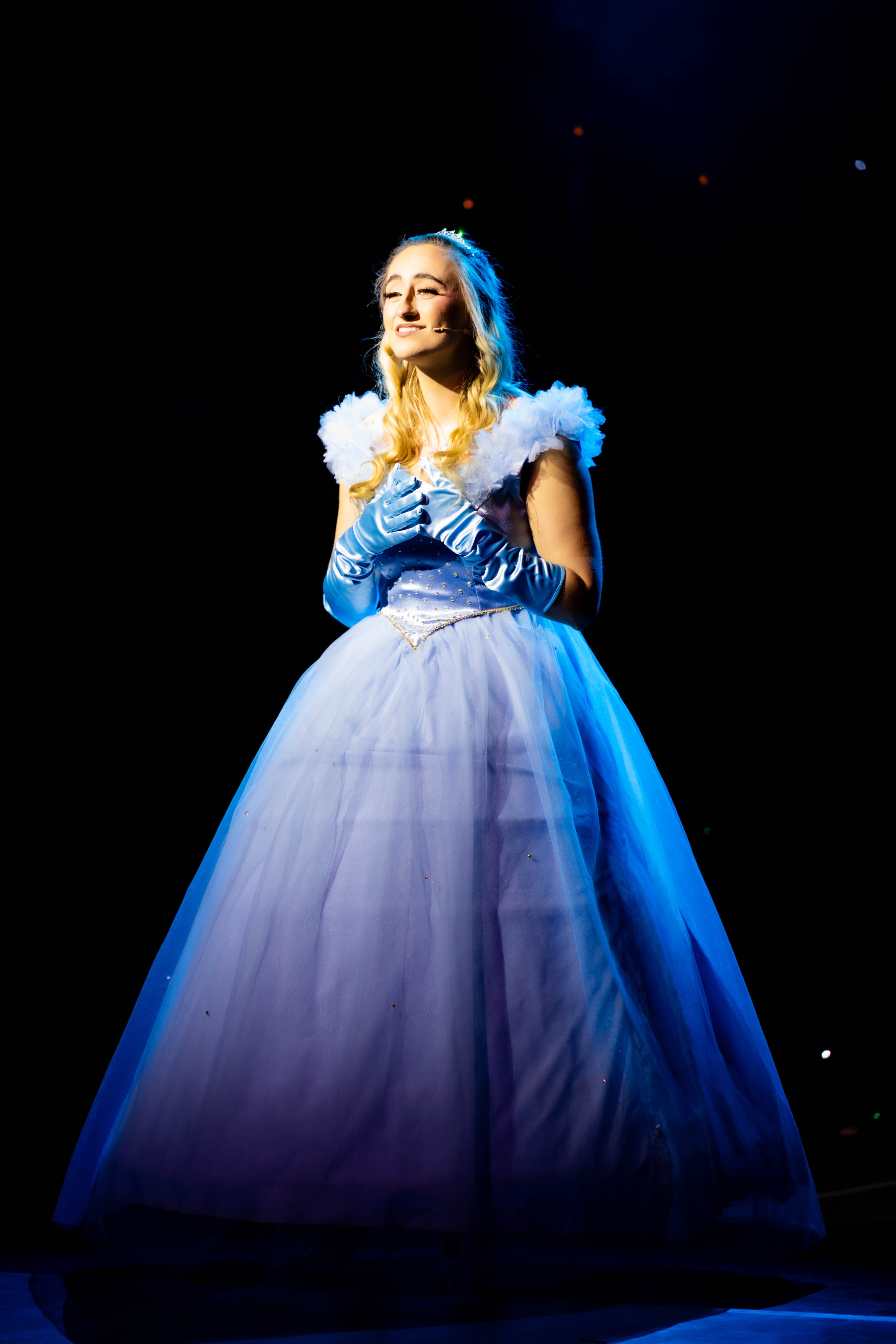 Cinderella poses with her hands on her heart while singing a solo. She wears a big ballroom dress. She is in a white spotlight, with the rest of the stage pitch black. Some stars sparkle behind her.